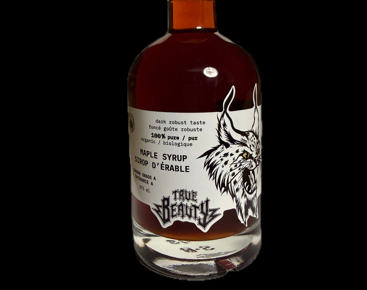 A bottle of True Beauty Dark Amber Maple Syrup against a solid black background. The bottle is made of clear glass showing the dark amber liquid inside, sealed with a black cap, and features a white label with a black graphic of a lynx head and the brand name 'True Beauty'.