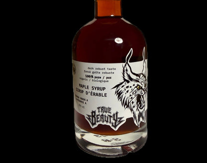 A bottle of True Beauty Dark Amber Maple Syrup against a solid black background. The bottle is made of clear glass showing the dark amber liquid inside, sealed with a black cap, and features a white label with a black graphic of a lynx head and the brand name 'True Beauty'.