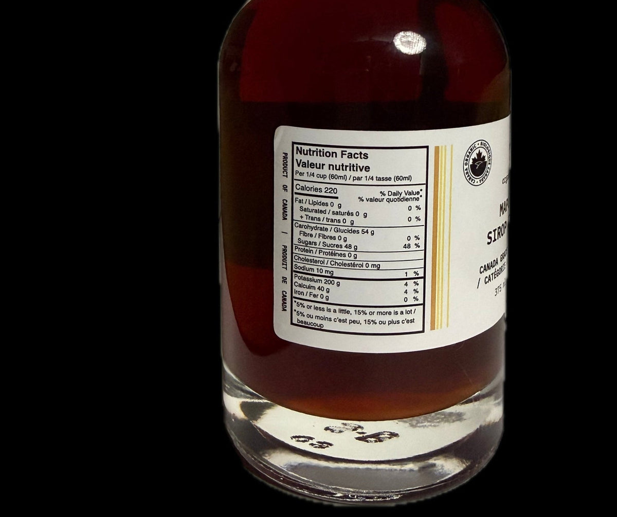 A clear glass bottle filled with amber-brown liquid maple syrup shown against a solid black background. The bottle has a black ribbed cap, a white nutrition label on the front, and a small circular logo on the right side of the label.