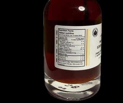 A clear glass bottle filled with amber-brown liquid maple syrup shown against a solid black background. The bottle has a black ribbed cap, a white nutrition label on the front, and a small circular logo on the right side of the label.